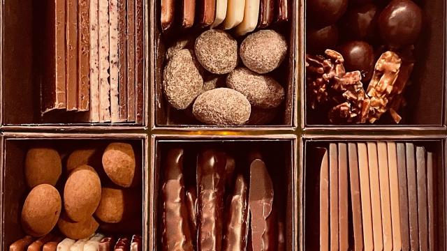 A close-up of an assorted chocolate box divided into six compartments, each holding multiple different types of chocolates: truffles, chocolate-dipped sticks, caramel clusters, and layered bars.
